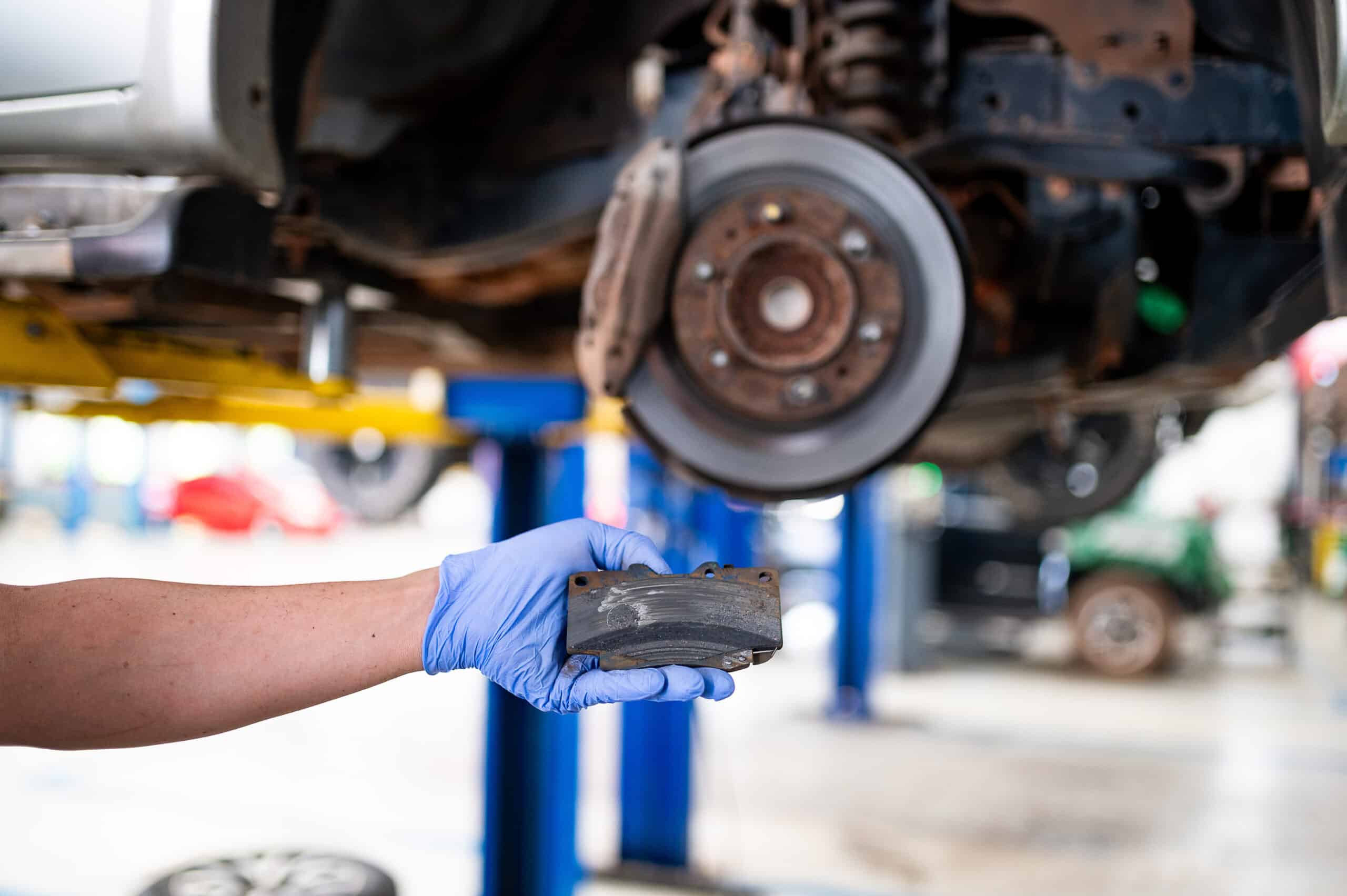 A gloved hand holds a worn brake pad in front of a raised car with an exposed brake disc at an auto repair shop.